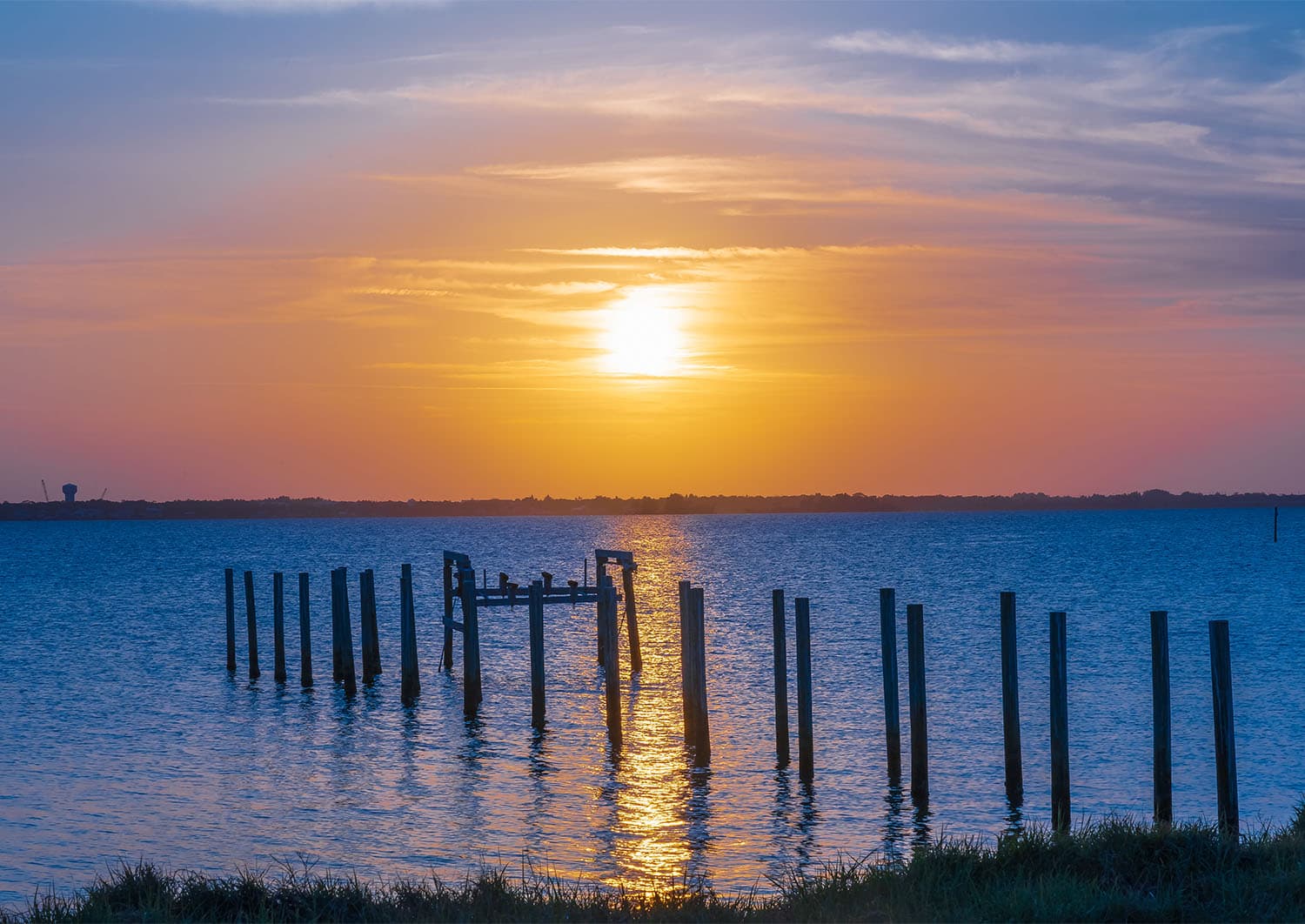 Sunset over the Indian River Lagoon with wooden dock pilings silhouetted against a golden sky, sunlight reflecting on calm waters