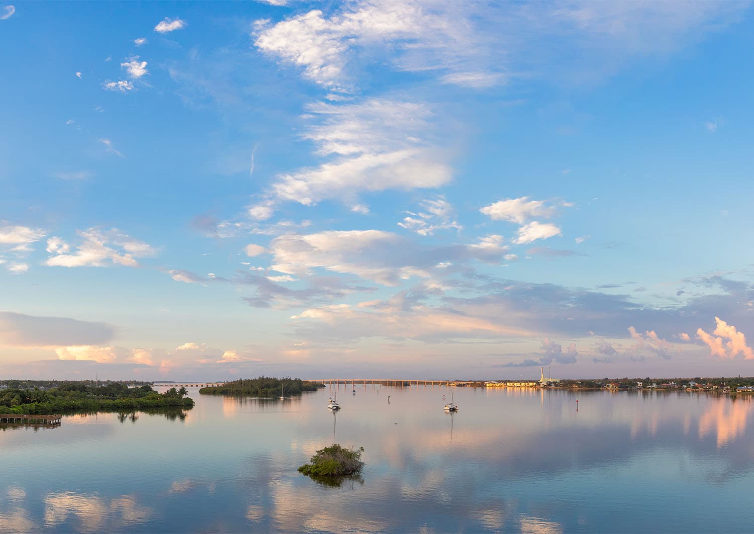 Panoramic view of the Indian River Lagoon at dawn with scattered clouds reflected in still waters, a bridge and anchored sailboats visible in the distance