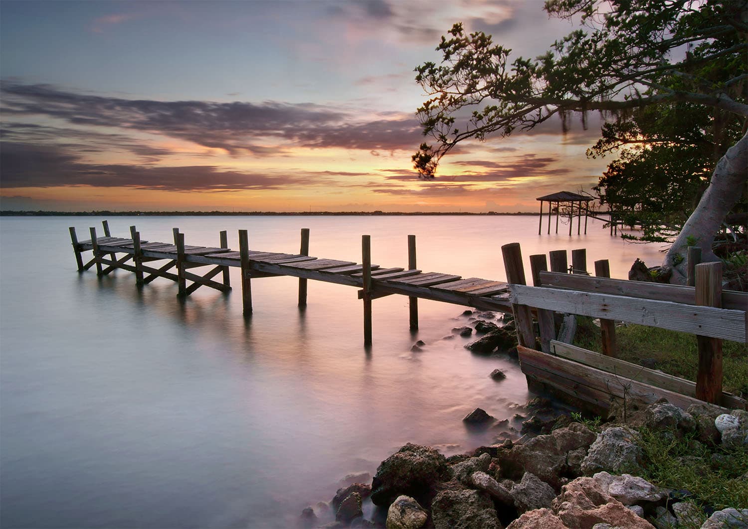 A weathered wooden dock extending into the Indian River Lagoon at sunset with rocks along the shoreline and overhanging tree branches